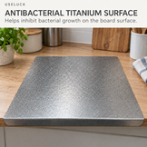 A square gray cutting board with rounded corners sits on a wooden kitchen countertop. Large text at the top reads 'ANTIBACTERIAL TITANIUM SURFACE' with a smaller 'USELUCK' mark above it, and kitchen items are softly blurred in the background.