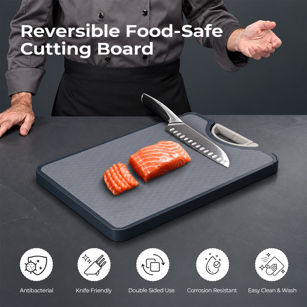 A dark gray rectangular cutting board with a black border is shown on a dark countertop, with a chef's hands and torso visible behind it. A large knife and a smaller paring knife rest on the board, along with two slices of salmon.