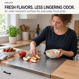 A woman in a navy long-sleeve shirt slices an apple on a gray silicone cutting board set into a light wood kitchen countertop. She holds a white bowl of apple slices.