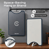A foldable cutting board shown in two states: hanging on a black wall rack on the left, and folded flat on a wooden table on the right. The product has a dark gray textured front, a white underside, and a navy blue border.