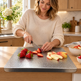 A woman in a cream knit sweater slices strawberries and apple slices on a speckled gray cutting board in a bright kitchen.